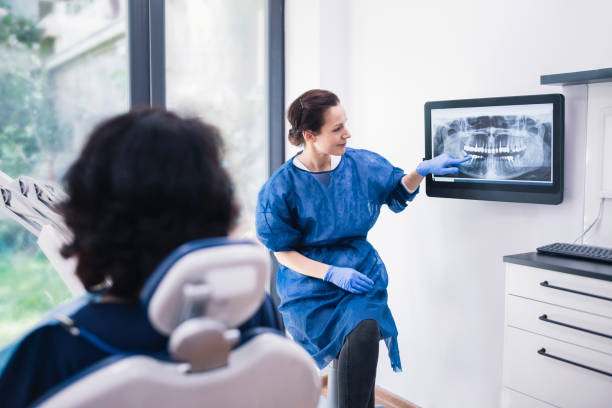 A dentist wearing blue scrubs and gloves points to a screen with a digital radiograph of a patient’s mouth. A patient sits in a chair facing her.