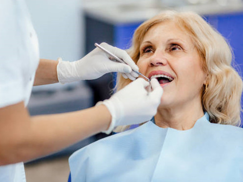 A woman sits in a chair smiling while a practitioner uses a dental tool in her mouth.